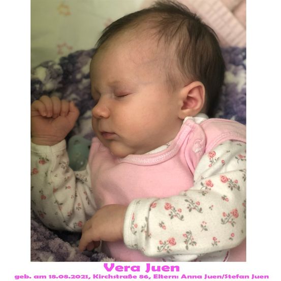 Ein Babymädchen mit braunem Haar schläft friedlich auf einer lila Decke. Sie trägt ein rosafarbenes, mit Blumen bedrucktes Lätzchen und ein langärmeliges Shirt. Das Bild hat einen Wasserzeichen mit Datum und Elternnamen.