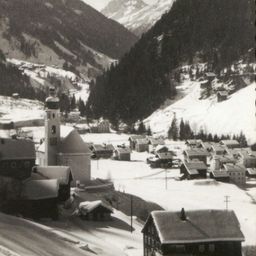 Eine Schwarz-Weiß-Fotografie eines Dorfes in den Bergen, mit einem Kirchturm und schneebedeckten Dächern.