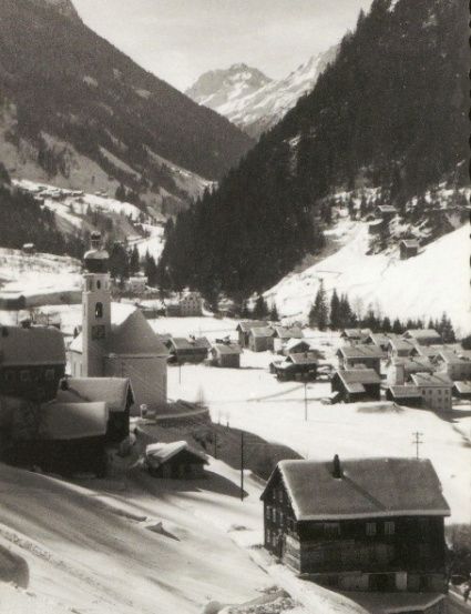 Eine Schwarz-Weiß-Fotografie eines Dorfes in den Bergen, mit einem Kirchturm und schneebedeckten Dächern.