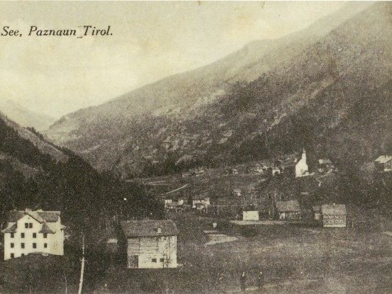 Ein altes Schwarz-Weiß-Foto zeigt eine bergige Landschaft mit einem Dorf im Vordergrund, das eine Kirche und mehrere Häuser umfasst.