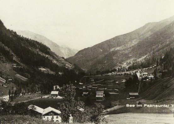 Ein altes Schwarz-Weiß-Foto eines gebirgigen Tals mit einem kleinen Dorf zwischen den Hügeln. Bäume und Häuser säumen die Landschaft.