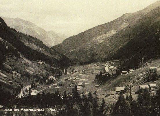 Ein altes Schwarz-Weiß-Foto zeigt ein Tal mit mehreren kleinen Häusern, die zwischen Bäumen und Bergen eingebettet sind. Das Tal ist weit und von Hügeln umgeben.