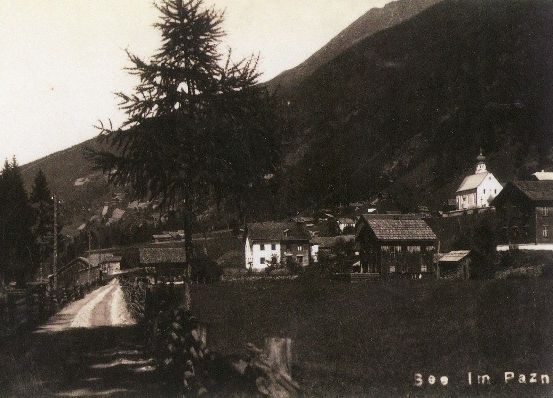 Ein sepiafarbenes Foto zeigt ein kleines Dorf in den Bergen, mit einer Kirche und mehreren Häusern. Ein Feldweg führt in das Dorf, umgeben von Bäumen und Bergen.