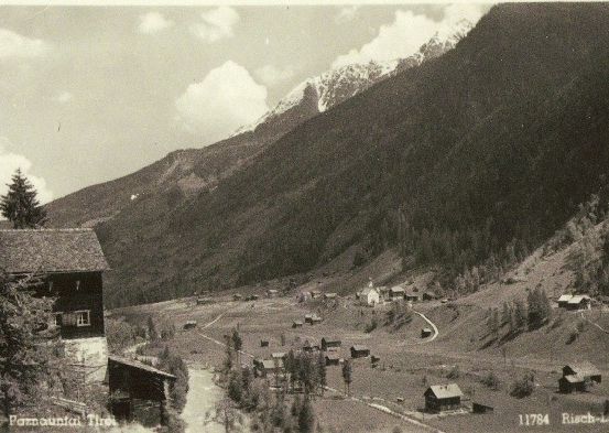 Ein Schwarz-Weiß-Foto eines Bergdorfes mit einem Fluss, der hindurchfließt. Das Dorf hat mehrere kleine Häuser und eine Kirche in der Ferne. Die Berge sind mit Bäumen und Schnee bedeckt.
