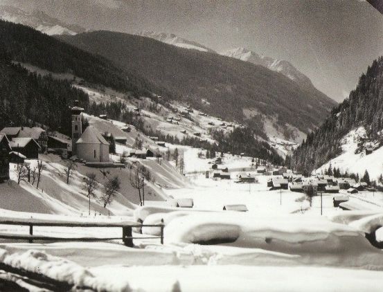 Ein Schwarz-Weiß-Foto eines verschneiten Bergdorfes mit Häusern und einer Kirche in der Ferne. Im Hintergrund sind Bäume und Berge zu sehen.