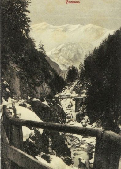 Ein altes Postkartenbild zeigt eine verschneite Berglandschaft mit einer Brücke und einem Holzzaun. Der Text lautet 'Trisannaschlucht mit dem Ausblick auf Wiesberg, Paznaun.'