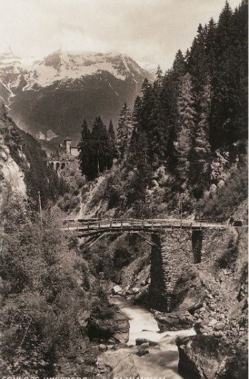 Ein Schwarz-Weiß-Foto einer Brücke über einem Fluss mit Bergen im Hintergrund.