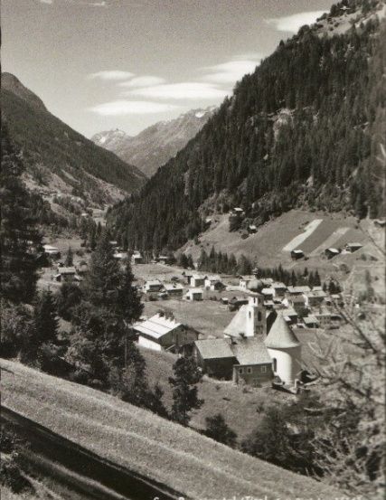 Ein Schwarz-Weiß-Foto eines kleinen Dorfes in den Bergen mit einer Kirche in der Mitte.