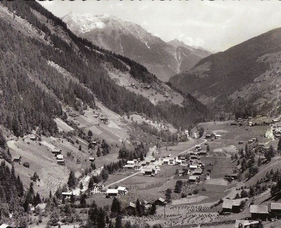 Eine Schwarz-Weiß-Luftaufnahme eines kleinen Dorfes in den Bergen, umgeben von dichten Wäldern und schneebedeckten Gipfeln.