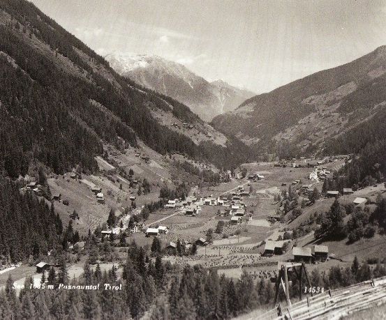 Eine Schwarz-Weiß-Luftaufnahme eines Tals mit einem kleinen Dorf, das zwischen den Bergen liegt. Das Dorf ist von Bäumen umgeben und hat einige Häuser. Die Berge sind schneebedeckt.