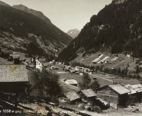 Ein Schwarz-Weiß-Foto eines kleinen Dorfes in den Schweizer Alpen, umgeben von üppigem Grün und hohen Bergen.