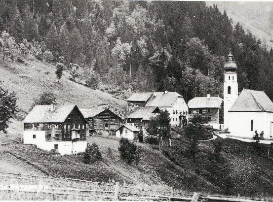 Ein Schwarz-Weiß-Foto eines kleinen Dorfes, eingebettet in einem bergigen Gebiet, mit einer Kirche mit Glockenturm und mehreren Häusern.