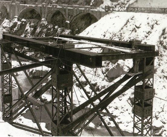 Ein Schwarz-Weiß-Foto einer Brückenbaustelle mit Stahlrahmen, Kabeln und einem verschneiten Berg im Hintergrund.