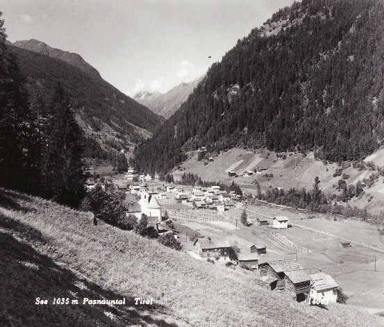 Ein Schwarz-Weiß-Foto eines kleinen Dorfes in den Bergen, mit einer Kirche im Zentrum. Bäume und Berge umgeben das Dorf, und ein Schild zeigt die Höhe an.