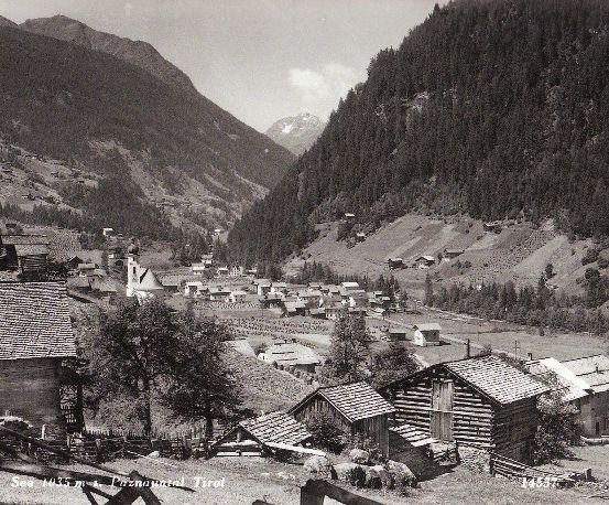 Ein Schwarz-Weiß-Foto eines kleinen Dorfes, eingebettet zwischen Bergen, mit traditionellen Holzhäusern und einer Kirche im Zentrum. Das Dorf ist von üppigem Grün und Bergen umgeben.