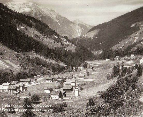 Eine Schwarz-Weiß-Fotografie eines Bergdorfes mit traditionellen Holzhäusern, umgeben von dichten Wäldern und schneebedeckten Bergen im Hintergrund.