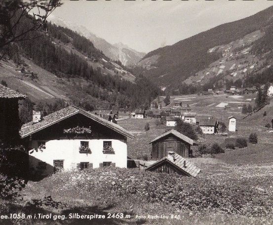 Ein Schwarz-Weiß-Foto eines Dorfes in den Bergen, mit mehreren Häusern und einer Kirche in der Ferne. Im Vordergrund befindet sich ein großes Haus mit einem Garten und Blumen.