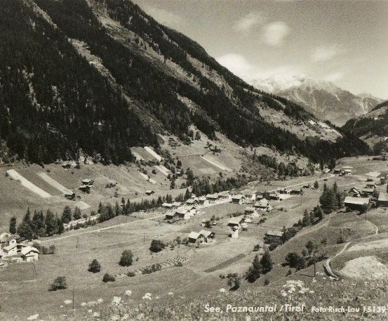 Ein Schwarz-Weiß-Foto eines kleinen Dorfes in den Bergen, umgeben von Bäumen und einer kurvenreichen Straße. Das Dorf befindet sich im Paznaun-Tal, Tirol.