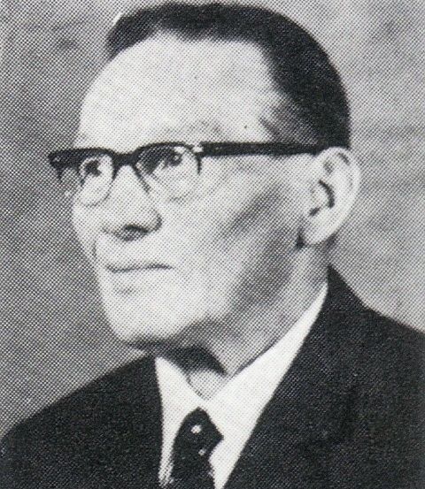 Schwarz-Weiß-Porträt eines Mannes in Anzug und Krawatte, mit Brille, mit ernster Miene.
