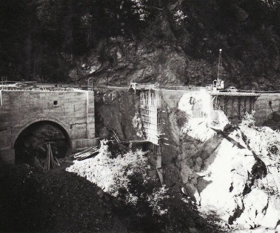 Ein Schwarz-Weiß-Foto zeigt eine Baustelle mit einer Brücke im Bau auf einem Berg. Die Brücke wird von Säulen gestützt, und es sind Fahrzeuge und Arbeiter zu sehen.