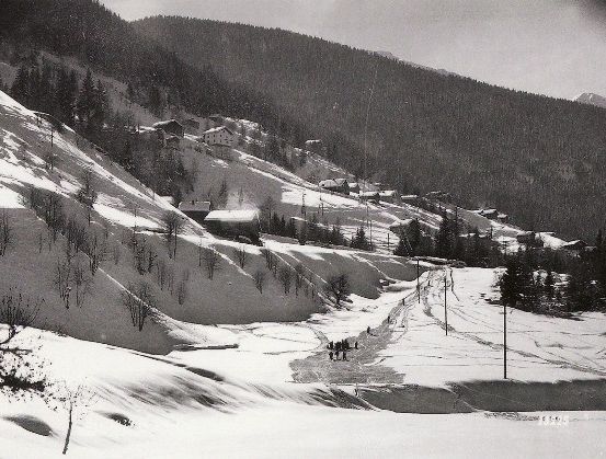 Ein Schwarz-Weiß-Foto einer verschneiten Berglandschaft mit mehreren Häusern im Tal. Eine Gruppe von Menschen geht auf einem Schneeweg.
