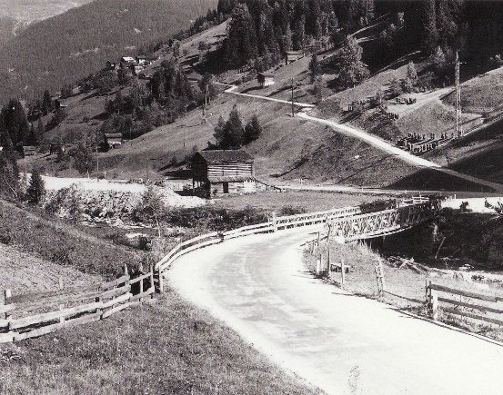 Ein Schwarz-Weiß-Foto zeigt eine kurvenreiche Bergstraße mit Holzzäunen auf beiden Seiten. Eine Holzbrücke überspannt die Straße, und ein kleines Holzhaus ist am Hang zu sehen.