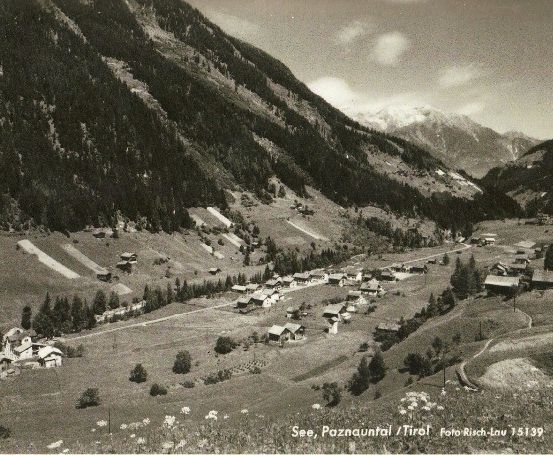 Ein Schwarz-Weiß-Foto eines bergigen Tals mit kleinen Häusern und einer kurvenreichen Straße. Das Tal ist von Bäumen und Bergen umgeben, mit einem schneebedeckten Gipfel in der Ferne.