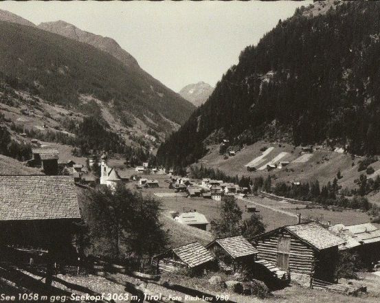 Ein Schwarz-Weiß-Foto eines kleinen Dorfes in einem Bergtal, mit Holzhäusern und einer kurvenreichen Straße.
