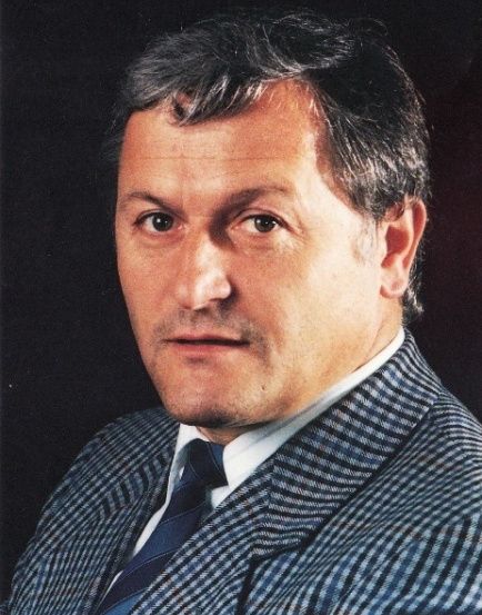 Ein Porträt eines mittelalten Mannes mit kurzen, dunklen Haaren, der eine blau-weiß karierte Anzugjacke und eine blaue Krawatte trägt. Er hat einen ernsten Gesichtsausdruck.