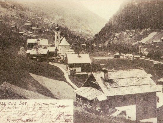Ein altes Schwarz-Weiß-Foto zeigt ein Bergdorf mit einer Kirche und mehreren Häusern. Das Bild enthält eine handgeschriebene Notiz auf Deutsch.