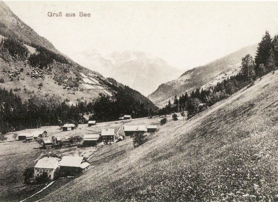 Eine Schwarz-Weiß-Fotografie eines kleinen Dorfes in den Bergen, mit einigen Häusern und einem Bergrücken im Hintergrund.