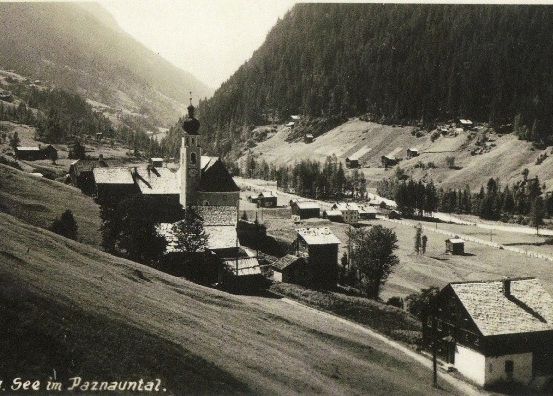 Ein Schwarz-Weiß-Foto eines Bergdorfs mit einer Kirche mit Turm und mehreren Häusern. Das Dorf liegt in einem Tal, umgeben von Bäumen und Bergen. Das Bild hat einen Wasserzeichen mit dem Text '1947 See im Paznauntal.'