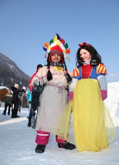 Zwei Frauen in Kostümen, eine mit einem bunten Kopfschmuck und die andere in einem gelben Kleid, stehen im Schnee mit Bergen im Hintergrund.