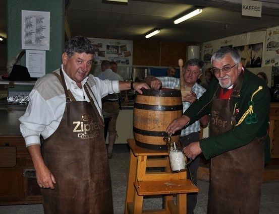 Zwei Männer in Schürzen gießen Bier in einen Holzfass in einer rustikalen Umgebung.