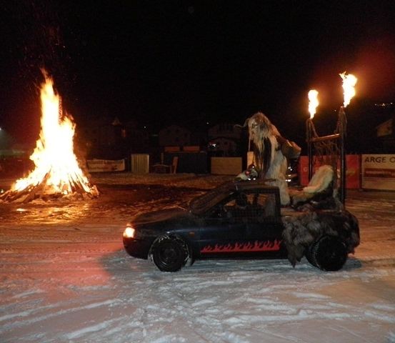 Ein Auto mit zwei brennenden Fackeln auf dem Dach steht nachts vor einem großen Lagerfeuer.