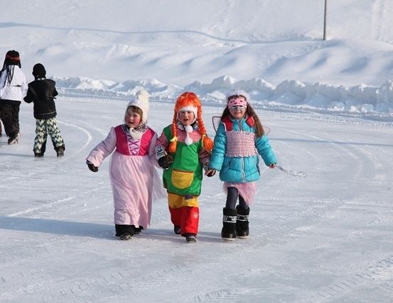 Drei Kinder in bunten Kostümen gehen auf einer verschneiten Oberfläche, mit zwei Erwachsenen hinter ihnen. Der Schnee ist an den Seiten aufgehäuft.