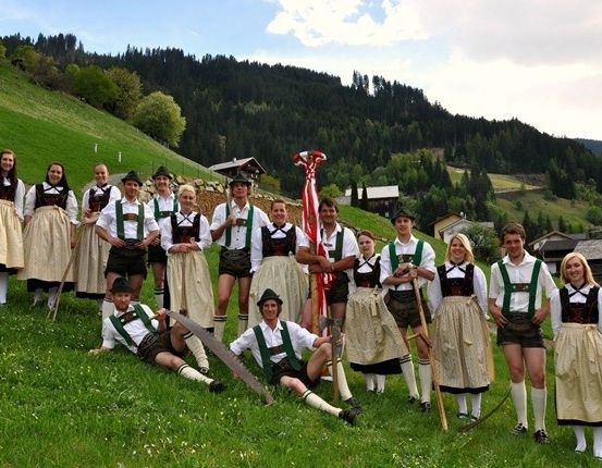 Eine Gruppe von Menschen in traditioneller bayerischer Kleidung posiert für ein Foto auf einem Grashügel mit Bergen im Hintergrund.