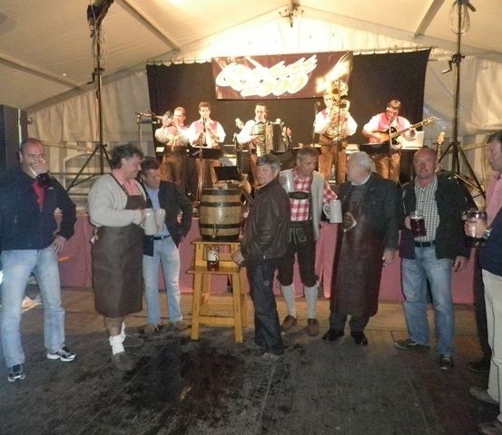 Eine Gruppe von Menschen versammelt sich in einem Zelt, während eine Band im Hintergrund spielt. Einige halten Getränke in den Händen, und ein Fass steht auf einem kleinen Tisch im Vordergrund.