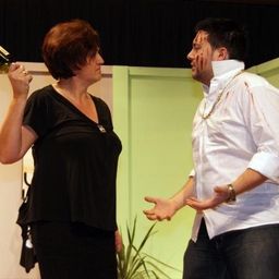 Eine Frau hält eine Flasche Wein und ein Mann mit Blut im Gesicht stehen sich gegenüber und schauen sich an.