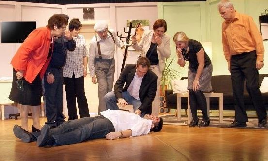 Eine Gruppe von Menschen versammelt sich in einem Raum, wobei eine Person auf dem Boden liegt. Die anderen schauen besorgt auf ihn herab. Der Raum hat eine Couch, einen Tisch und einen Fernseher an der Wand.