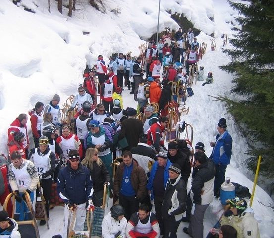 Eine Gruppe von Menschen in Winterkleidung steht in einem verschneiten Bereich, einige halten Schlitten. Sie scheinen auf ein Schlittenrennen zu warten.