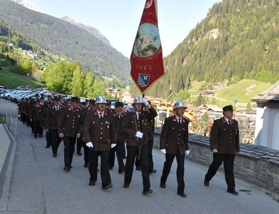Eine Gruppe uniformierter Personen, die bei einer Parade mit einer Flagge, auf der das Jahr 1982 steht, durchzieht, vor dem Hintergrund eines Gebirgsgeländes und eines Steingebäudes.