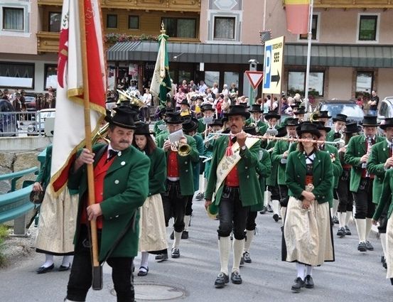 Eine Musikkapelle in grünen und weißen Uniformen, die Flaggen und Musikinstrumente tragen, tritt bei einem Umzug auf einer Stadtstraße auf.
