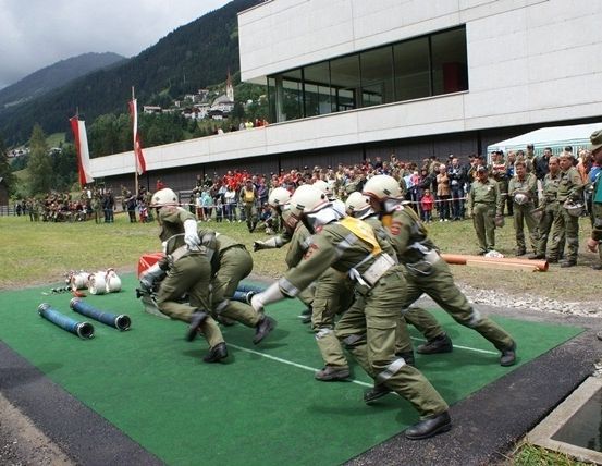 Eine Gruppe Feuerwehrleute in Uniform übt eine Übung auf einem grünen Teppich vor einem Gebäude, das von einer Menge beobachtet wird.