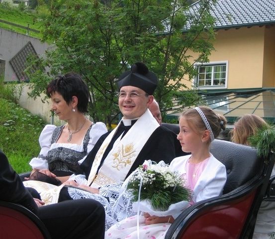 Ein Priester und ein junges Mädchen in einem Blumenkleid sitzen in einer Kutsche mit einer Frau und einem Mann. Sie befinden sich in einer ländlichen Umgebung mit einem Haus und einem Baum im Hintergrund.