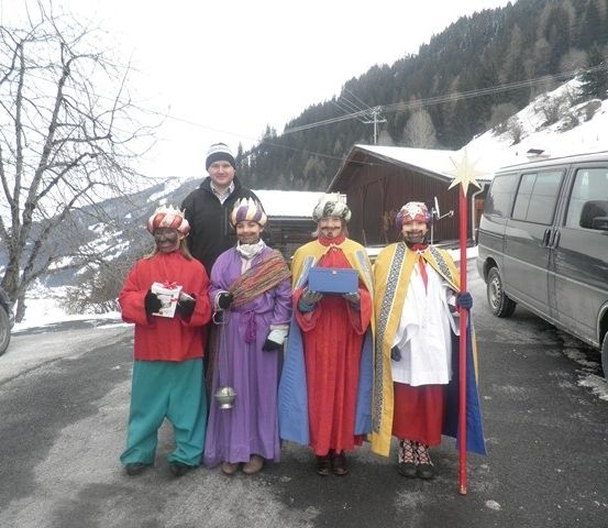 Fünf Personen in farbenfrohen Kostümen, möglicherweise für einen festlichen Anlass, stehen in einem verschneiten Bereich mit einem Van und einem Haus im Hintergrund.