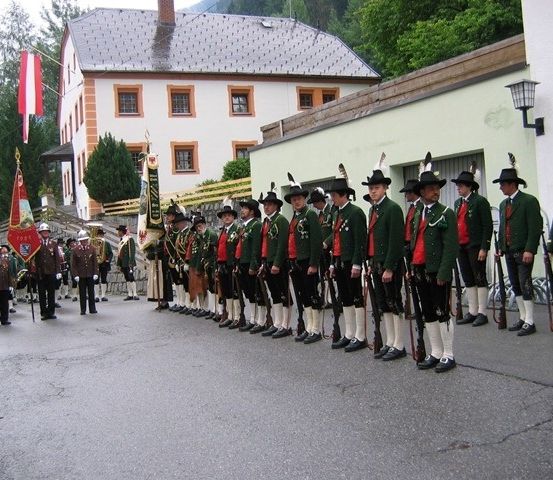 Eine Gruppe von Männern in grünen Uniformen und Hüten steht in einer Reihe und hält Gewehre, möglicherweise für eine Parade vor einem Gebäude.
