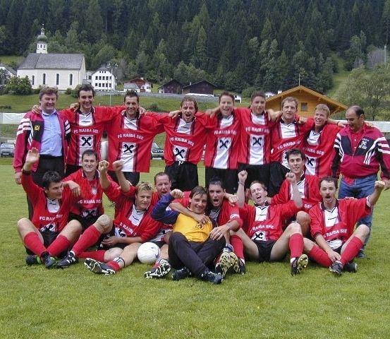 Eine Fußballmannschaft in roten und weißen Trikots posiert für ein Gruppenfoto auf einem Rasenfeld mit Häusern und Bäumen im Hintergrund.