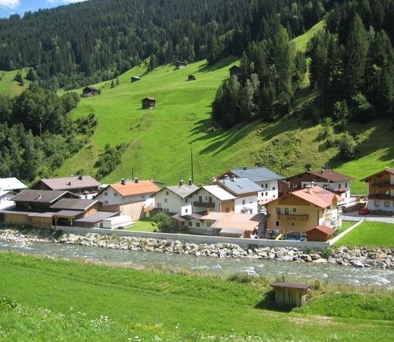 Ein malerisches Dorf in den österreichischen Alpen, umgeben von üppigen grünen Wiesen und dichten Wäldern, mit einem kleinen Fluss, der hindurchfließt.