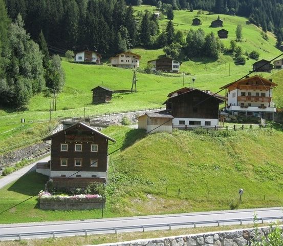 Luftaufnahme eines kleinen Dorfes in den österreichischen Alpen, mit traditionellen Holzhäusern, üppigen grünen Wiesen und einer kurvenreichen Straße.